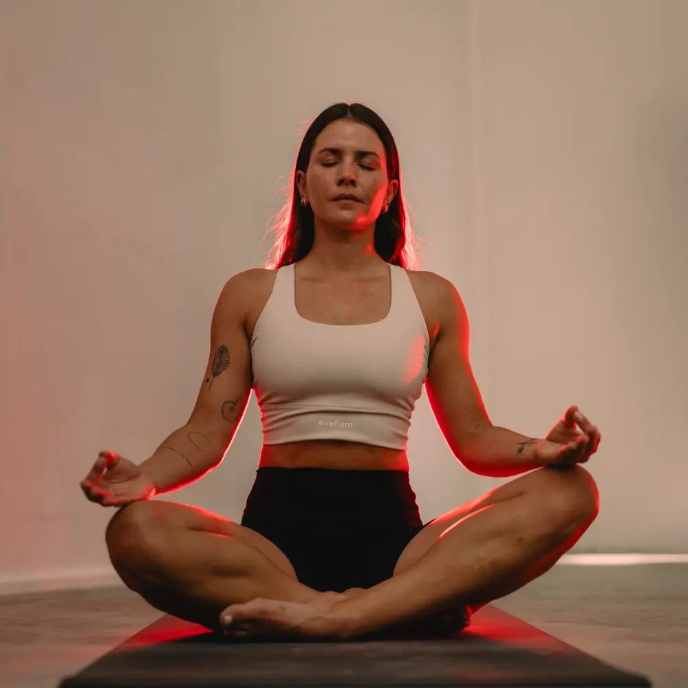 Frontalansicht einer meditierenden Frau im Lotussitz auf einer Yoga-Matte, die von hinten durch das Reboots Relight Pro Rotlicht angestrahlt wird.