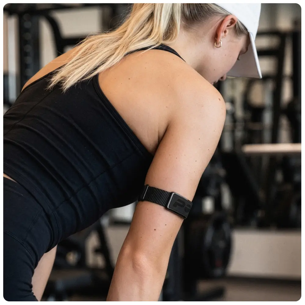 Lifestyle-Foto einer Sportlerin in schwarzer Kleidung im Fitnessstudio. Sie trägt den POLAR Loop Fitness Tracker mit der bildschirmlosen Vorderseite nach außen am Oberarm fixiert, gehalten von einem dunkelgrauen Sportarmband aus flexiblem Gewebe.