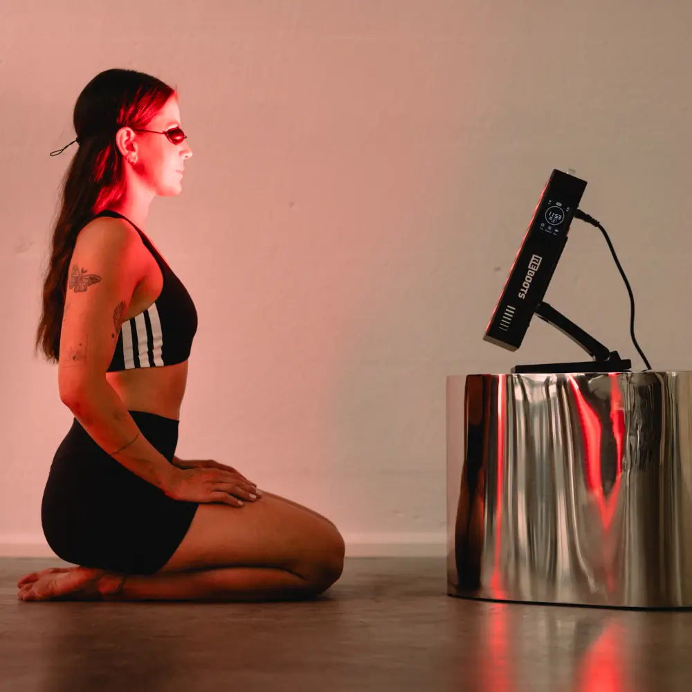 Eine Sportlerin mit Laserschutzbrille kniet vor dem eingeschalteten Reboots Relight Mini Rotlichttherapie-Panel für eine Oberkörperanwendung.