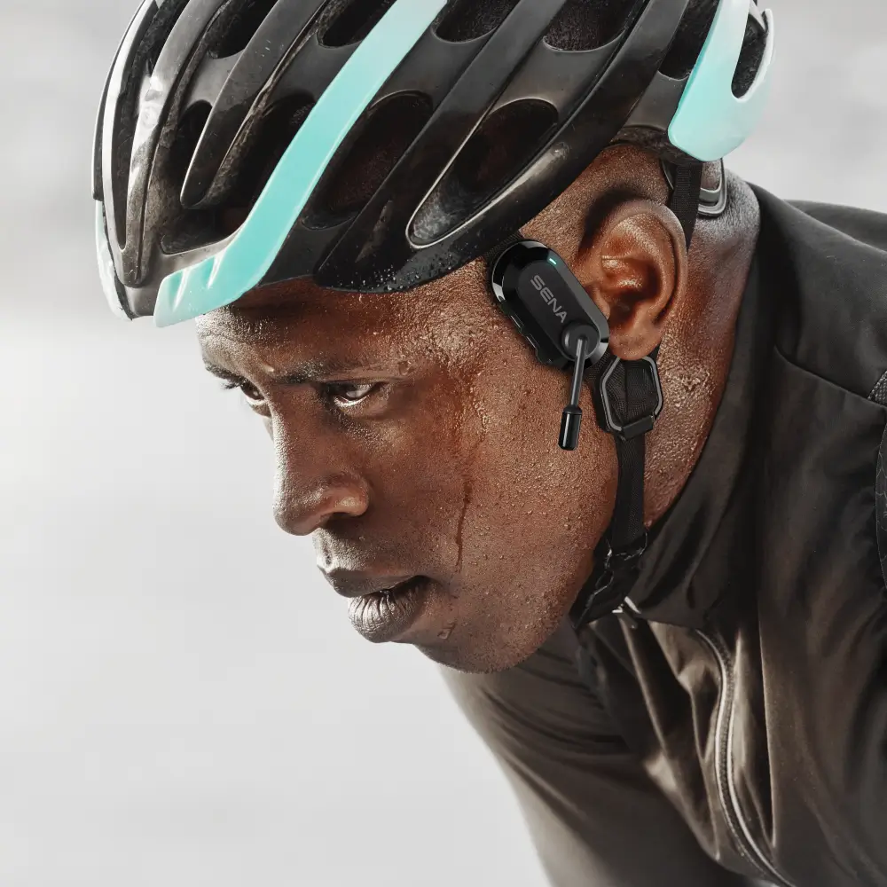 Nahaufnahme eines intensiv trainierenden, verschwitzten Radfahrers mit schwarzem Helm. Am Helmriemen auf Ohrhöhe ist das schwarze Sena BiKom 20 Kommunikationssystem befestigt, dessen Mikrofon nach vorne gerichtet ist.